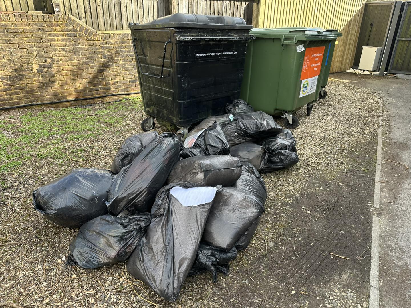 A pile of black garbage bags left on the ground next to two large waste bins in an outdoor area.