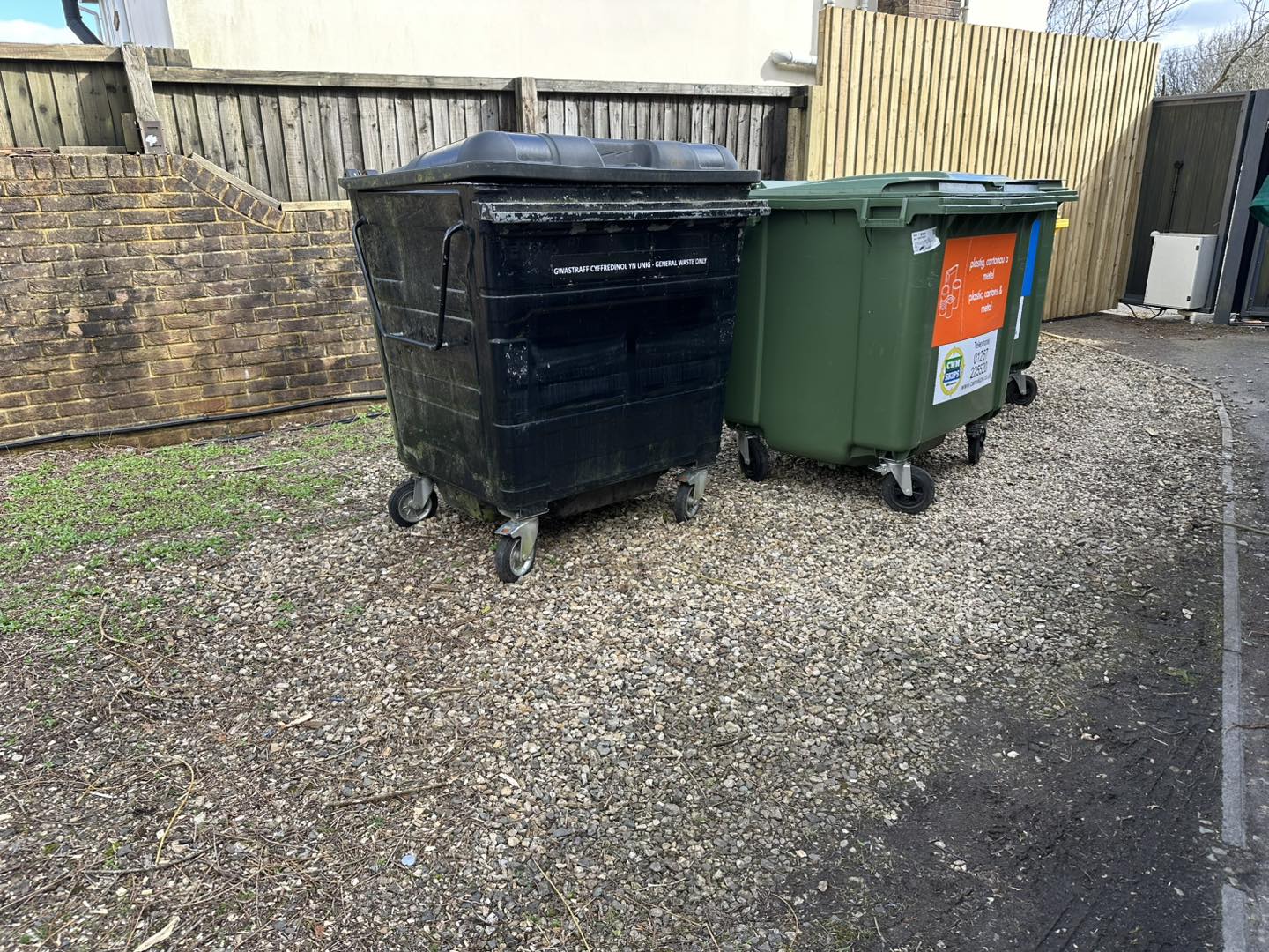 Two large outdoor waste bins, one black and one green, stand on a gravel area next to a wooden fence.